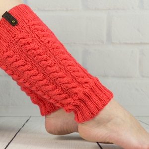 Puede incluir: Un par de polainas de punto de color coral con un patrón de punto trenzado. Las polainas tienen una etiqueta de cuero marrón con un botón de metal.