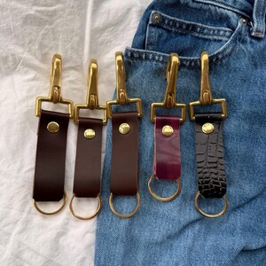 May include: Five leather keyrings with brass hooks and rings. The keyrings are in different colours and textures, including brown, burgundy, and black crocodile embossed leather.