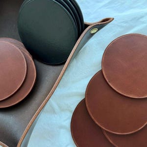Puede incluir: Un juego de posavasos de cuero marrón y un juego de posavasos de cuero negro en una bandeja de cuero marrón.