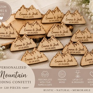 May include: A collection of light-colored wooden mountain-shaped cutouts, each engraved with the text Emma & Josh and the date 07.19.2025. These small, flat decorative pieces feature a jagged peak design and are crafted from natural wood.