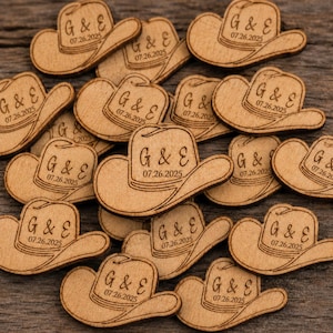 May include: A collection of small, light brown wooden cutouts shaped like cowboy hats. Each hat features the engraved text G & E and the date 07.26.2025. The wooden pieces are scattered across the frame, displaying a natural wood grain texture.