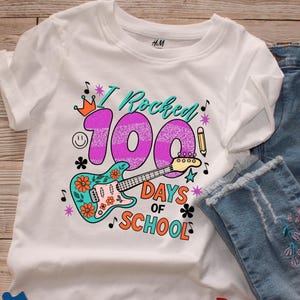 May include: White t-shirt with a colorful graphic celebrating 100 days of school. The design includes a guitar, the text "I Rocked 100 Days of School", musical notes, and a crown.
