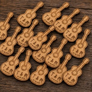 May include: A collection of small, light brown wooden guitar-shaped cutouts. Each piece features the engraved text T & M and the date 07.26.2025. The guitar shapes have a circular sound hole and a long neck, arranged in a scattered pattern.