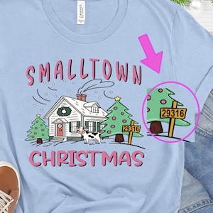 May include: Pale blue t-shirt with a festive graphic. The design includes a house, Christmas trees, a cow, and the number 29316. The words "Smalltown Christmas" are in pink, with green and black accents.