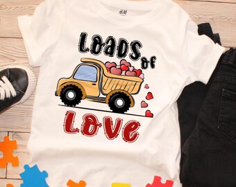 May include: White t-shirt with a graphic of a yellow dump truck carrying heart-shaped cargo. The text "Loads of Love" is printed in black and red. Black jeans and a sneaker are visible.