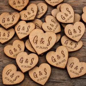 Può includere: Una collezione di piccoli pezzi di legno a forma di cuore, ognuno inciso con le lettere G & S e la data 07.26.2025. Il legno marrone chiaro mostra una venatura naturale e il testo è impresso sulla superficie in una tonalità marrone scuro.