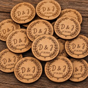 Può includere: Una collezione di dischi circolari in legno chiaro, ognuno inciso con un bordo decorativo a corona d'alloro che circonda le iniziali D & J e la data 07.26.2025. I pezzi di legno sono disposti in gruppo, mettendo in risalto la venatura naturale.