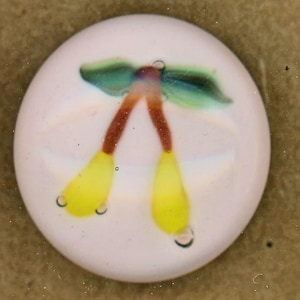 May include: A round, white button with a hand-painted fruit design. The fruit features yellow teardrop shapes, brown stems, and green leaves. The word "FRUIT" is written below the button.