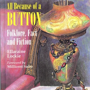 May include: A book cover with a colorful illustration of a lamp with a button necklace. The title of the book is "All Because of a BUTTON Folklore, Fact and Fiction" by Ellaraine Lockie. The book cover also includes the text "Foreword by Millicent Safro".