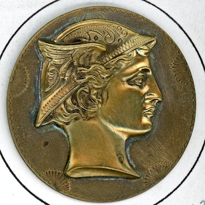 May include: A circular bronze-toned metal medallion featuring a relief profile of a figure wearing a winged helmet. The background of the medallion has a textured border with small fan-shaped patterns, and the central figure faces to the right.