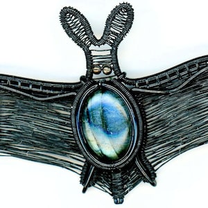 May include: A bat-shaped brooch crafted from dark wire, featuring an oval-shaped iridescent blue and grey gemstone at its center. The wirework forms the detailed wings and ears of the bat, creating a structured, layered appearance with a metallic finish.