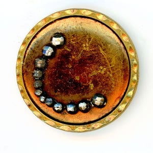 May include: A circular brass-toned button featuring a textured, scalloped edge and a weathered, copper-colored center. A curved row of small, faceted, dark metallic beads is embedded into the surface, creating a crescent-shaped accent.