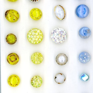 May include: A collection of decorative buttons in various shapes, sizes, and colors. The buttons include yellow, blue, and iridescent options, with designs such as floral, geometric, and textured patterns. Some buttons have gold or silver rims.