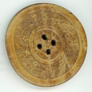 May include: A large, round, brown button with a floral design and four holes. The button is made of a natural material, possibly bone or wood.