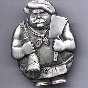 May include: A silver-toned metal pin shaped like a stout chef wearing a toque and neckerchief. The figure holds a large meat cleaver in one hand and a leg of meat in the other, with a textured apron over a shirt, rendered in a relief style.