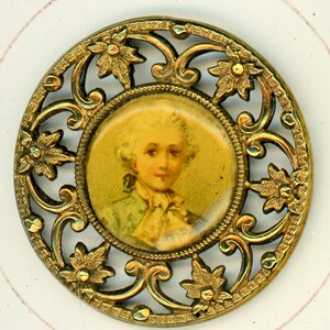 May include: A gold-toned metal button with a decorative filigree design. The button features a round, glass-like center with a portrait of a young person with a powdered wig.