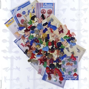 May include: A collection of colorful plastic buttons in various shapes and sizes, including animals, people, and objects. The buttons are arranged on a white background with a blue and white striped pattern. The text "SMILE! WE ARE LOOKING AT REALISTIC SETS" is at the top of the image. The text "National Button Society December 2024" is at the bottom of the image. The text "Ann Olson" is in the bottom right corner of the image.