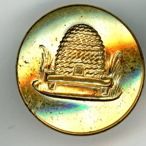 May include: A circular gold-toned button featuring a raised relief design of a skep beehive resting on a stand, surrounded by stylized grass. The surface of the button has a reflective, iridescent finish with subtle rainbow-colored highlights.