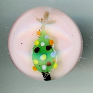 May include: A glass bauble with a pink background and a green Christmas tree with white and yellow spots. The tree has a gold star on top.