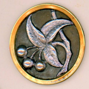 May include: A silver and gold button with a raised design of a leafy branch with berries. The button has a circular shape and a gold rim.