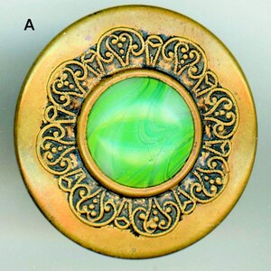 May include: A round, antique-style button with a brass-colored metal frame. The button features an ornate, dark-brown filigree design around a central, circular insert of swirled green glass. The letter "A" is visible in the upper left corner.