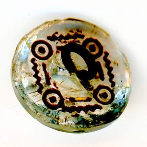 May include: A clear glass button with a brown design. The design features a circle with a smaller circle inside, surrounded by a wavy line. There are three of these circles on the button.