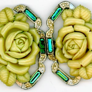 May include: A vintage, two-piece buckle featuring pale yellow rose and leaf designs. The metalwork is silver-toned, accented with clear and emerald green rhinestones. The buckle is likely for a belt or other garment.