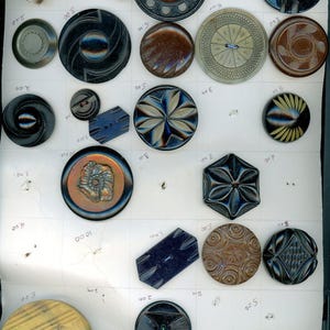 May include: A collection of vintage buttons in various shapes, sizes, and colors, including black, brown, and iridescent hues. Some buttons feature intricate designs, such as floral patterns and geometric shapes. The buttons are displayed on a white surface.