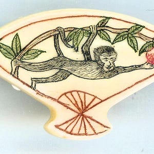 May include: A fan-shaped decorative object in an off-white tone featuring a detailed black ink illustration of a monkey hanging from a branch with green leaves and reaching toward a small red fruit, with a geometric fan pattern at the base.
