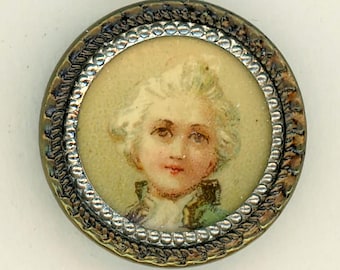 May include: A round, silver-toned button with a raised, beaded border. The button features a painted portrait of a young person with white hair, wearing a green and white jacket.