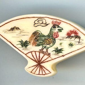May include: A fan-shaped brooch with an off-white base featuring a hand-drawn illustration of a green rooster standing on a fan design. The scene includes a red sun, a brown mountain, a cloud, and green grass accents in brown ink.