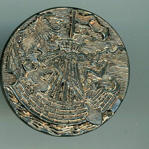 May include: A circular, silver-colored button with a raised design of a ship with a flag and a dragon. The button has a textured surface.