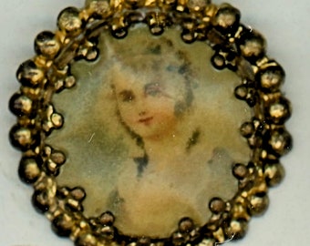 May include: A small, round antique brooch with a portrait of a woman. The portrait is surrounded by a gold-colored beaded frame. The woman in the portrait has light skin and is wearing a white dress.