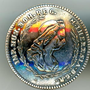 May include: A silver coin featuring a profile of a woman with a draped head covering, surrounded by inscriptions and a laurel wreath. The coin has a detailed edge and a slightly iridescent surface, with hints of red, yellow, and blue.