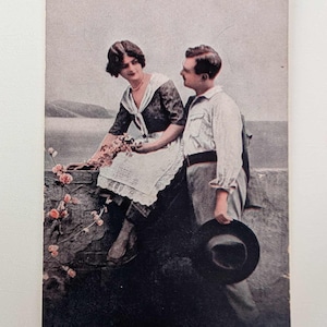 Postal romántica antigua de una pareja junto al mar. Postal coloreada a mano de la década de 1910. Pareja de enamorados junto al mar con flores.