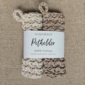May include: Two rectangular crocheted potholders in cream and tan colors, each featuring a small loop at the top. A white paper band wraps around the center with the text: HANDMADE Potholder 100% Cotton machine wash gentle - dryer low.
