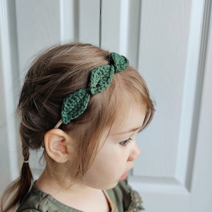 May include: A green crocheted headband featuring three leaf-shaped accents. The headband is worn by a young child with light brown hair and a green top. The background is a white door.