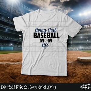 May include: White t-shirt with the text "living that BASEBALL MOM life" in navy blue and black. The design includes a baseball graphic. The shirt is displayed on a baseball field background.