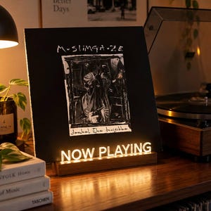 May include: A black vinyl record sleeve titled Muslimgauze Jackal The Invisible displayed on a wooden stand with a glowing white LED sign that reads NOW PLAYING. The stand is positioned on a wooden surface next to a stack of books and a record player.