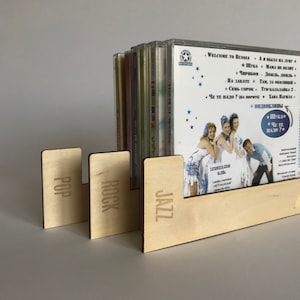 May include: A wooden CD rack with three sections labelled "Pop", "Rock", and "Jazz". The rack is holding a stack of CDs with Russian language text and images.