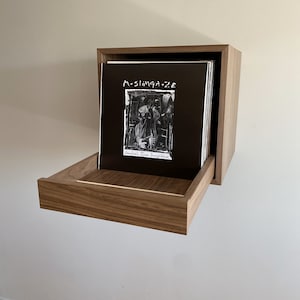 Puede incluir: Una estantería de pared de madera para discos de vinilo con un cajón. Un disco de vinilo está en la estantería con el título "Muslinga - 2E" y el texto "Jankbal the Invisible".