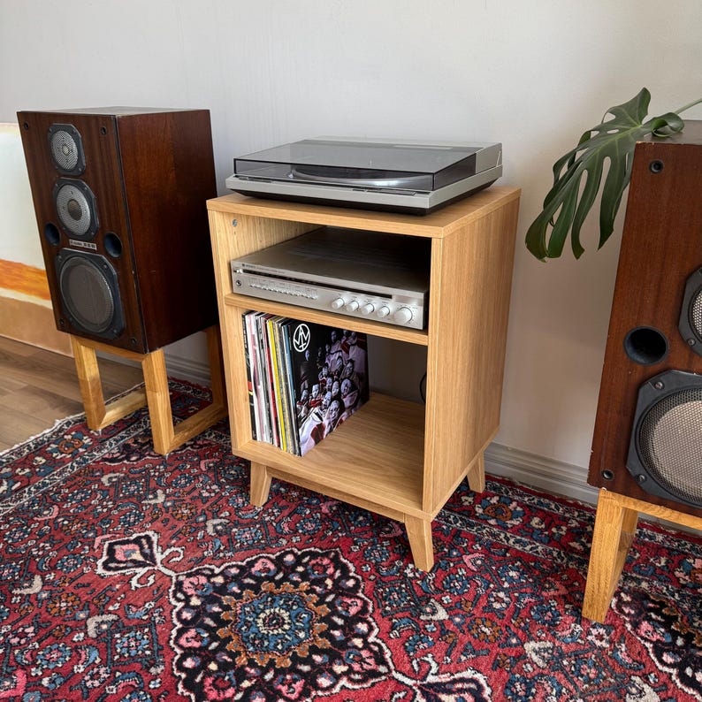 Puede incluir: Un mueble para tocadiscos de roble claro con un tocadiscos encima, un receptor plateado en el estante central y discos de vinilo almacenados verticalmente en el estante inferior, flanqueado por dos altavoces de madera oscura.