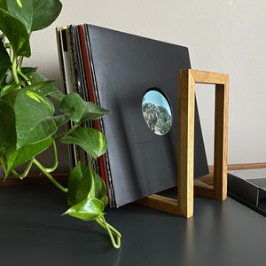 May include: A wooden record stand with a stack of black vinyl records leaning against it. The stand is made of light brown wood and has a simple, rectangular design.