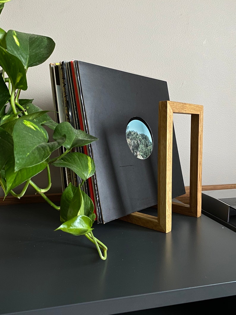 May include: A wooden record stand with a stack of black vinyl records leaning against it. The stand is made of light brown wood and has a simple, rectangular design.