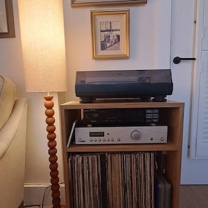 Puede incluir: Un mueble de madera para tocadiscos con un tocadiscos negro en la parte superior, un receptor de audio negro y plateado en el estante central y una colecci&oacute;n de discos de vinilo almacenados verticalmente en el estante inferior, junto a una l&aacute;mpara de pie.