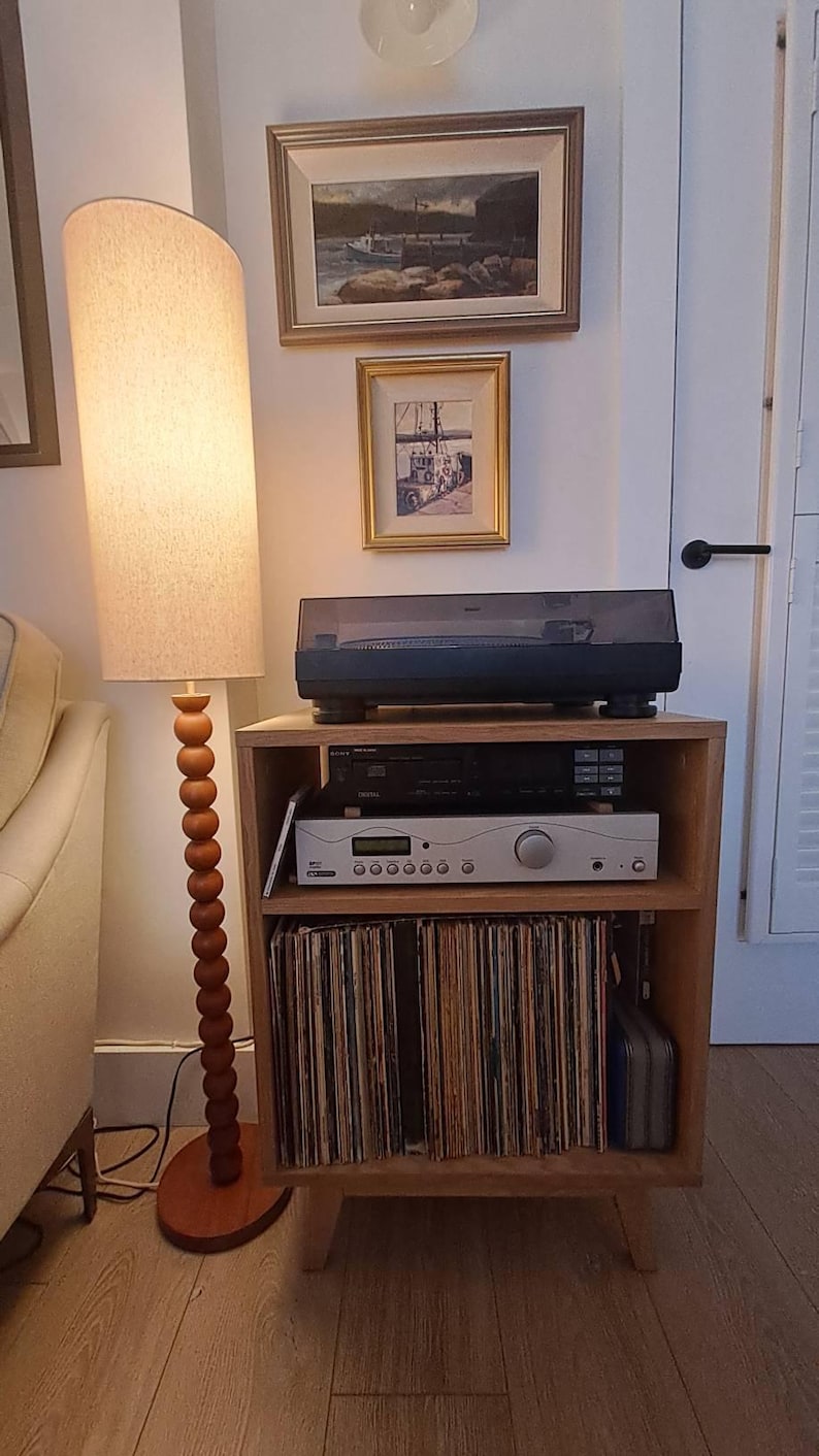 Puede incluir: Un mueble de madera para tocadiscos con un tocadiscos negro en la parte superior, un receptor de audio negro y plateado en el estante central y una colecci&oacute;n de discos de vinilo almacenados verticalmente en el estante inferior, junto a una l&aacute;mpara de pie.