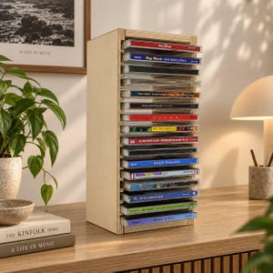 May include: A vertical light-colored wooden storage rack designed to hold compact discs. The unit features multiple horizontal shelves stacked to accommodate a collection of CD cases, each with visible spine text and colorful album cover art.