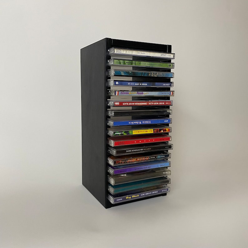 Peut inclure: Une tour de rangement noire pour CD avec 17 fentes, chacune pouvant accueillir un bo&icirc;tier de CD. La tour est remplie de CD, certains avec des pochettes color&eacute;es et d'autres avec des pochettes noires. Les CD sont &eacute;tiquet&eacute;s avec des titres et des artistes.