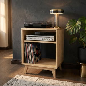 Puede incluir: Un mueble multimedia de madera de roble claro con cuatro patas c&oacute;nicas, que cuenta con un estante superior abierto con un tocadiscos negro, un estante central con un receptor de audio plateado y un estante inferior con discos de vinilo.