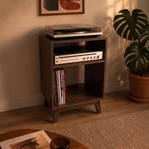 Puede incluir: Un soporte para tocadiscos de madera oscura con cuatro patas c&oacute;nicas, que cuenta con una superficie superior abierta para el tocadiscos, un estante central con un receptor de audio plateado y un compartimento inferior para discos.
